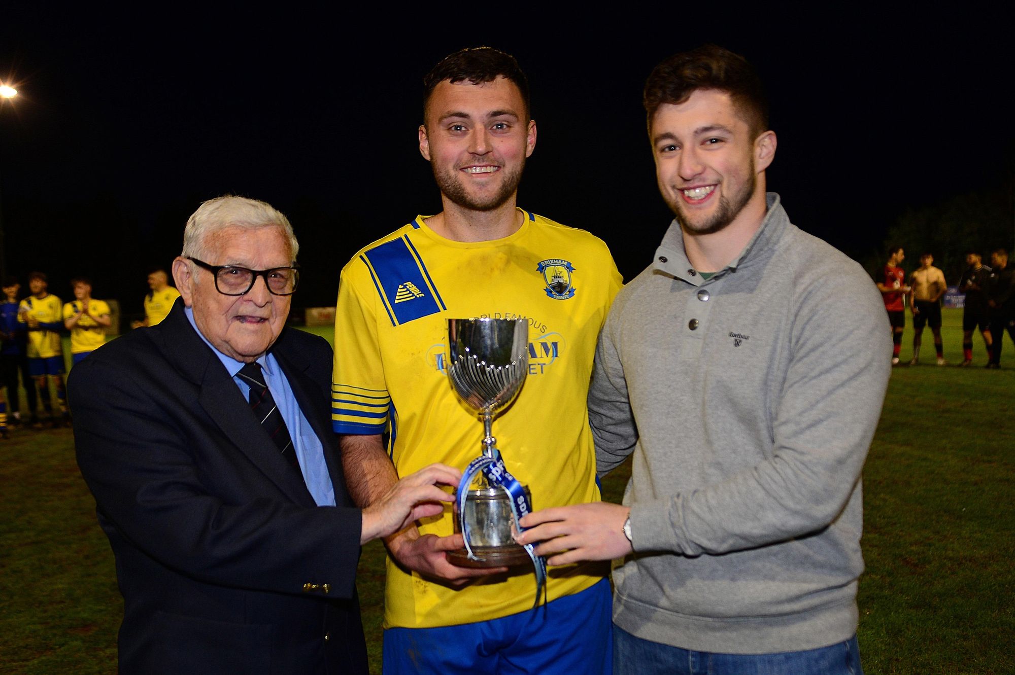 Captain Lewis Booth celebrates Brixham Town's Lidstone Cup final victory with SDFL president Mervyn Benney and sponsor George Helmore, of Bettesworths. Credit Al Macphee Miracle PR